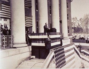 President Roosevelt stands at a podium surrounded by american flags at the top of the steps of a colonial brick building. 
